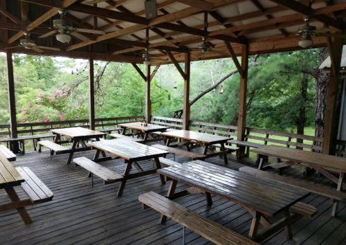 A spacious wooden pavilion with multiple picnic tables, surrounded by greenery and trees.