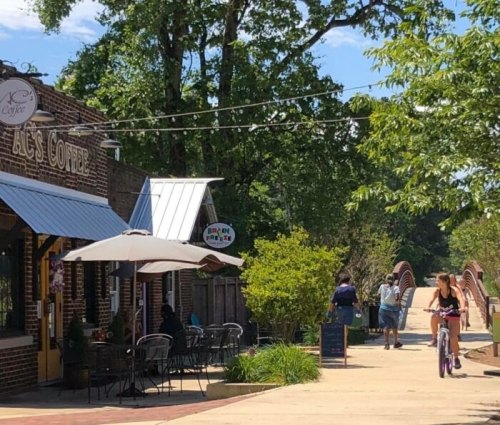 A sunny street scene with a coffee shop, outdoor seating, and people walking and biking along a path.
