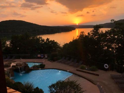Sunset over a tranquil lake, with a pool and lounge chairs in the foreground, surrounded by trees and hills.