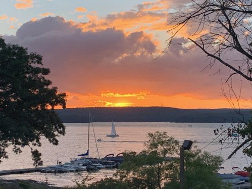 A serene sunset over a lake, with sailboats and silhouettes of trees in the foreground.