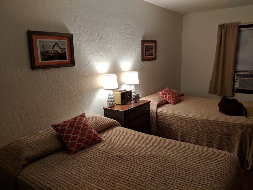A cozy bedroom with two beds, bedside lamps, and framed pictures on the wall, featuring warm lighting and a simple decor.