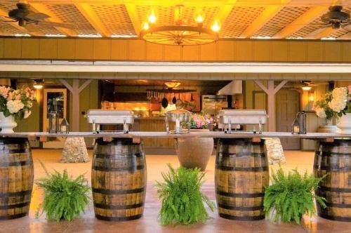 A rustic outdoor dining setup with wooden barrels, floral arrangements, and a kitchen in the background.