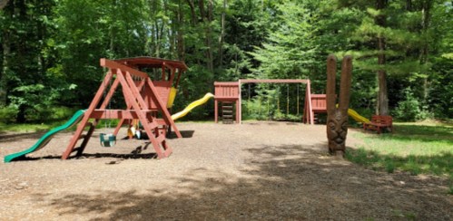 A playground with a slide, swings, and a wooden totem in a wooded area.