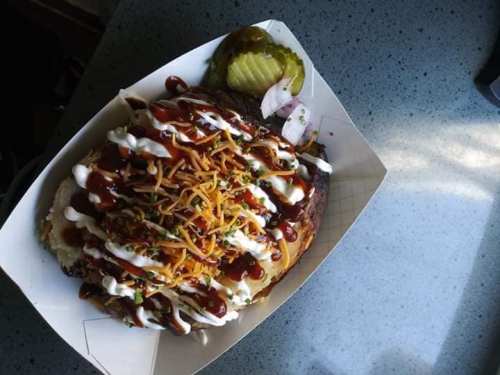 A loaded barbecue dish topped with cheese, sauce, onions, and pickles, served in a paper tray on a countertop.
