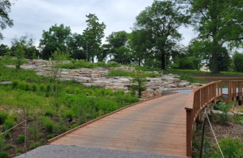 A wooden bridge leads through a landscaped area with rocks and greenery under a cloudy sky.