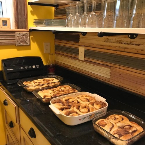 Freshly baked cinnamon rolls in glass dishes on a black countertop, with glasses and plates on a yellow wall shelf above.