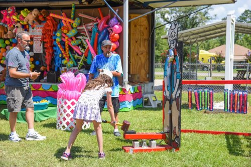 A girl plays a carnival game, hitting a target with a mallet while two adults watch. Colorful decorations in the background.