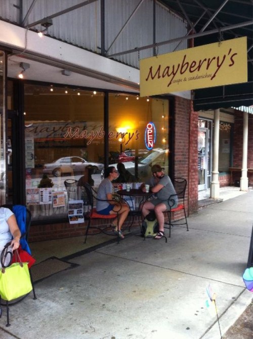 Two people sit at a small outdoor table outside Mayberry's cafe, enjoying food and drinks on a sunny day.