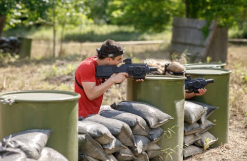 Two individuals in tactical gear aim laser tag guns from behind sandbag barriers in an outdoor setting.