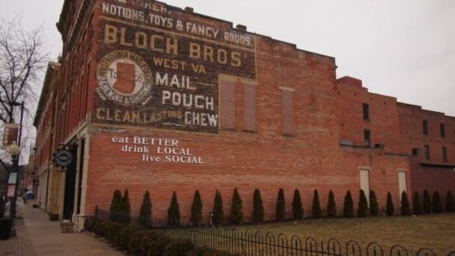 Faded mural on a brick building advertising Bloch Bros. with a grassy area and fence in front. Overcast sky.