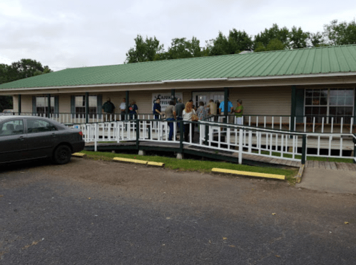 A group of people stands outside a building with a green roof, waiting near a ramp and parked cars.