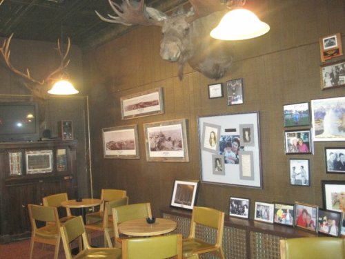 A cozy room with wooden walls, featuring moose heads, framed photos, and small tables with chairs.