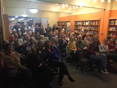 A large group of people sitting in a bookstore, holding up signs and smiling at the camera.