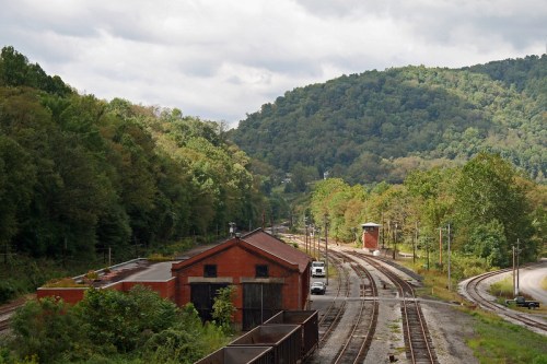 A scenic view of a railway yard with red brick buildings, tracks, and lush green hills in the background under a cloudy sky.