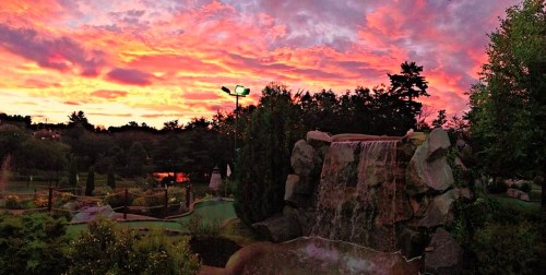A vibrant sunset with pink and orange hues over a mini-golf course featuring a waterfall and lush greenery.