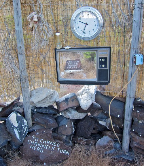 A vintage TV and clock mounted on a rustic wall, with a sign reading "Religion Channel for Sale" and a teddy bear.