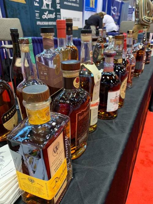 A display of various whiskey bottles lined up on a table at an event, with a blurred background of attendees.
