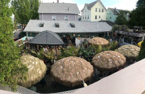A lively outdoor bar scene with thatched umbrellas, surrounded by greenery and colorful buildings in the background.