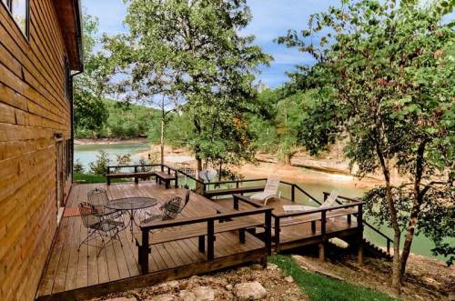 A wooden deck with chairs overlooks a serene lake surrounded by trees and greenery.