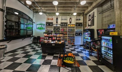 A modern bodega interior with checkered floors, shelves of snacks, drinks, and a display of fresh fruits.