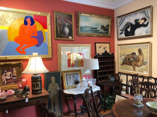 A cozy gallery filled with various framed artworks, a lamp, and antique furniture against a warm-colored wall.