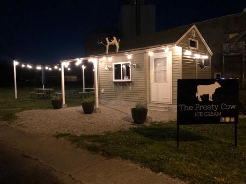 A small ice cream stand at night, illuminated by string lights, with a sign reading "The Frosty Cow Ice Cream."