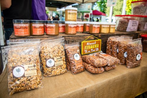 A market stall displaying jars of almond butter and bags of raw walnuts, surrounded by a vibrant outdoor setting.