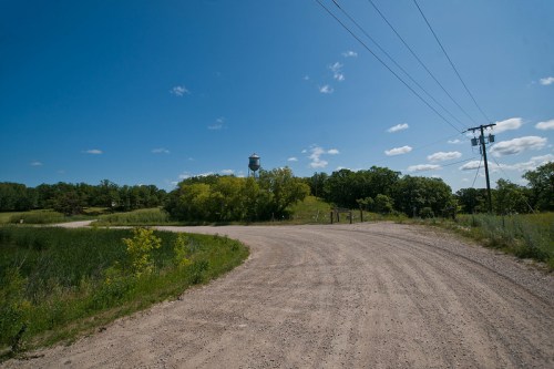 A winding dirt road leads to a water tower, surrounded by greenery and under a clear blue sky with scattered clouds.