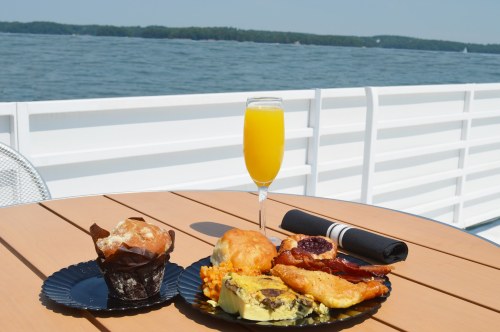 A plate of breakfast items including a muffin, quiche, bacon, and a mimosa, set against a scenic waterfront view.
