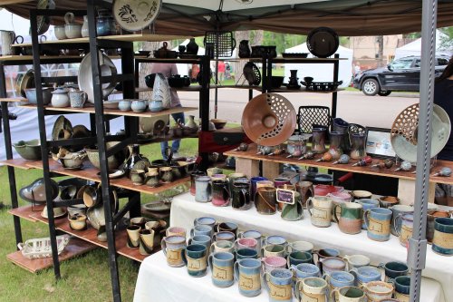 A display of handcrafted pottery, including mugs and decorative pieces, arranged on wooden shelves at an outdoor market.