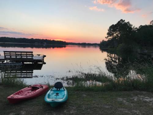 Two kayaks sit on the shore of a calm lake at sunset, with a dock and trees in the background.