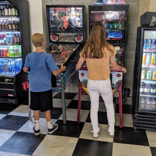 Two children play pinball at an arcade, focused on their games, with vending machines in the background.