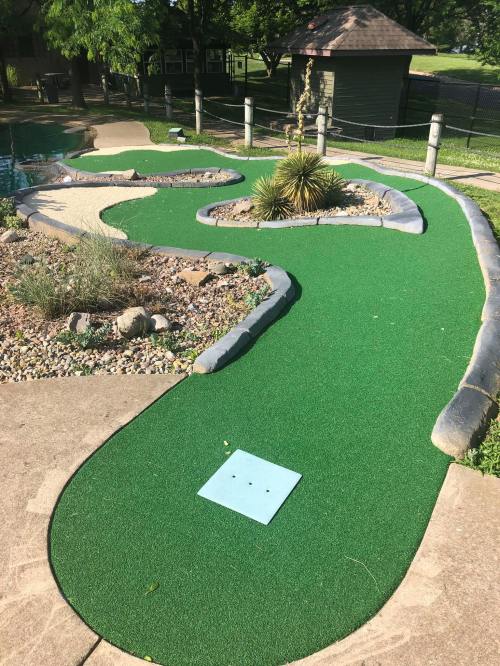 A mini-golf course featuring a winding green path, rocks, and a small plant, set in a sunny outdoor area.