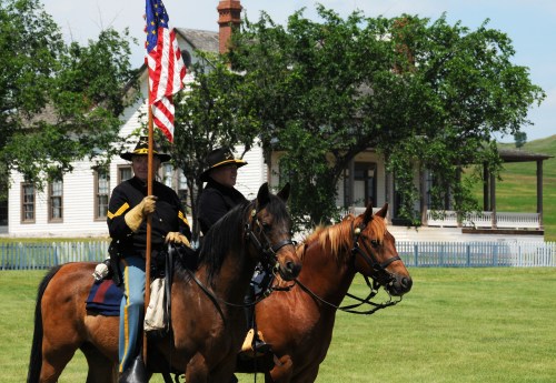 Two mounted soldiers in historical uniforms hold an American flag, with a house and trees in the background.