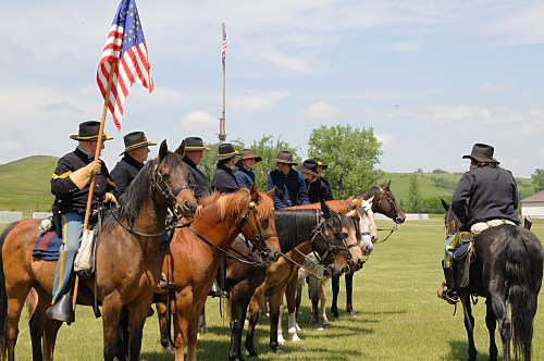 A group of mounted soldiers in historical uniforms, holding a U.S. flag, gathered on a grassy field.