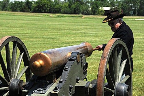 A man in a black coat and hat stands beside a historic cannon on a grassy field.