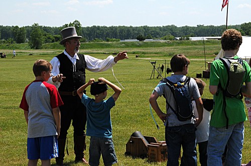 A man in period costume engages with a group of children outdoors on a sunny day, with tents in the background.