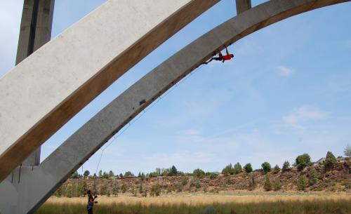 A person rappels down a concrete arch while another person stands below, set against a blue sky and grassy landscape.