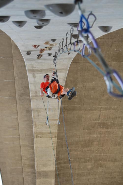 A climber in an orange shirt hangs from a rock climbing wall under a bridge, secured by ropes and harnesses.