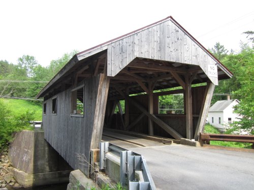A rustic wooden covered bridge spans a small road, surrounded by greenery and a few buildings in the background.
