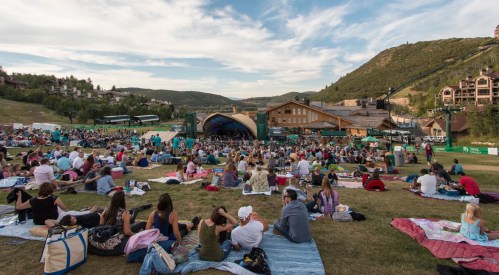 A large crowd enjoys a concert on a grassy field, with mountains and buildings in the background under a blue sky.