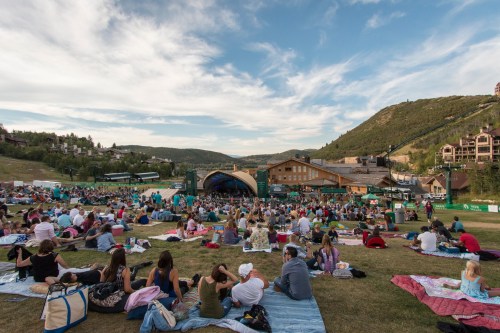 A large crowd sits on blankets on a grassy field, enjoying a concert at an outdoor venue surrounded by hills.