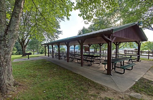 A shaded picnic shelter with multiple tables, surrounded by trees and a grassy area near a lake.
