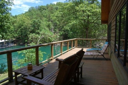 A wooden deck overlooking a serene lake, surrounded by lush green trees and a clear blue sky.