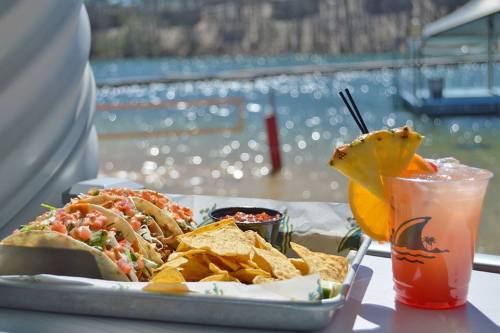 A plate of tacos with salsa and chips, alongside a colorful drink, with a scenic waterfront in the background.