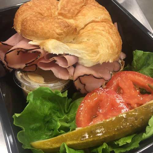 A croissant sandwich filled with ham, served with lettuce, tomato slices, a pickle, and a small cup of sauce.