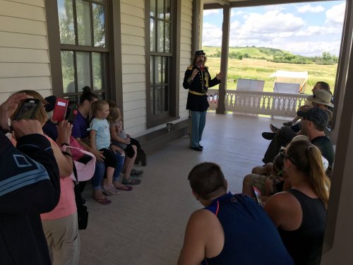 A historical reenactor in uniform speaks to a group of people seated on a porch, surrounded by a scenic landscape.
