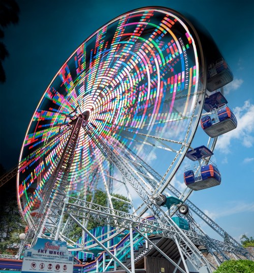 A colorful ferris wheel spins against a blue sky, with vibrant lights illuminating its structure.