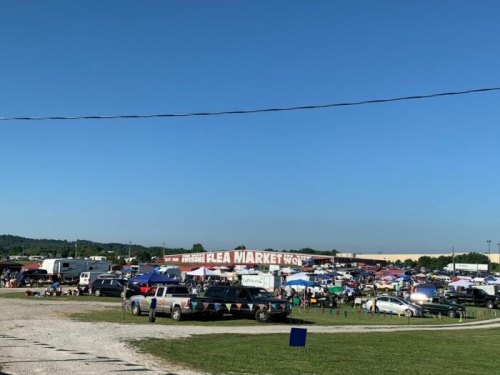 A bustling flea market with numerous vendors, cars, and shoppers under a clear blue sky.