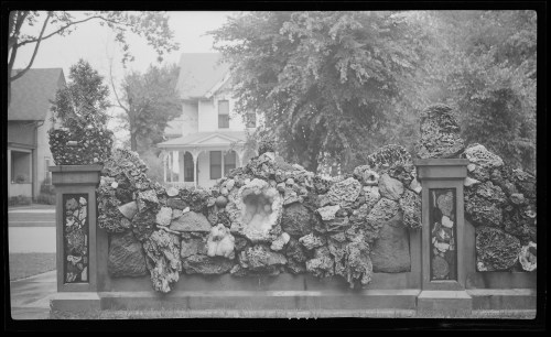A decorative stone wall with various rocks and plants, set against a backdrop of a house and trees.